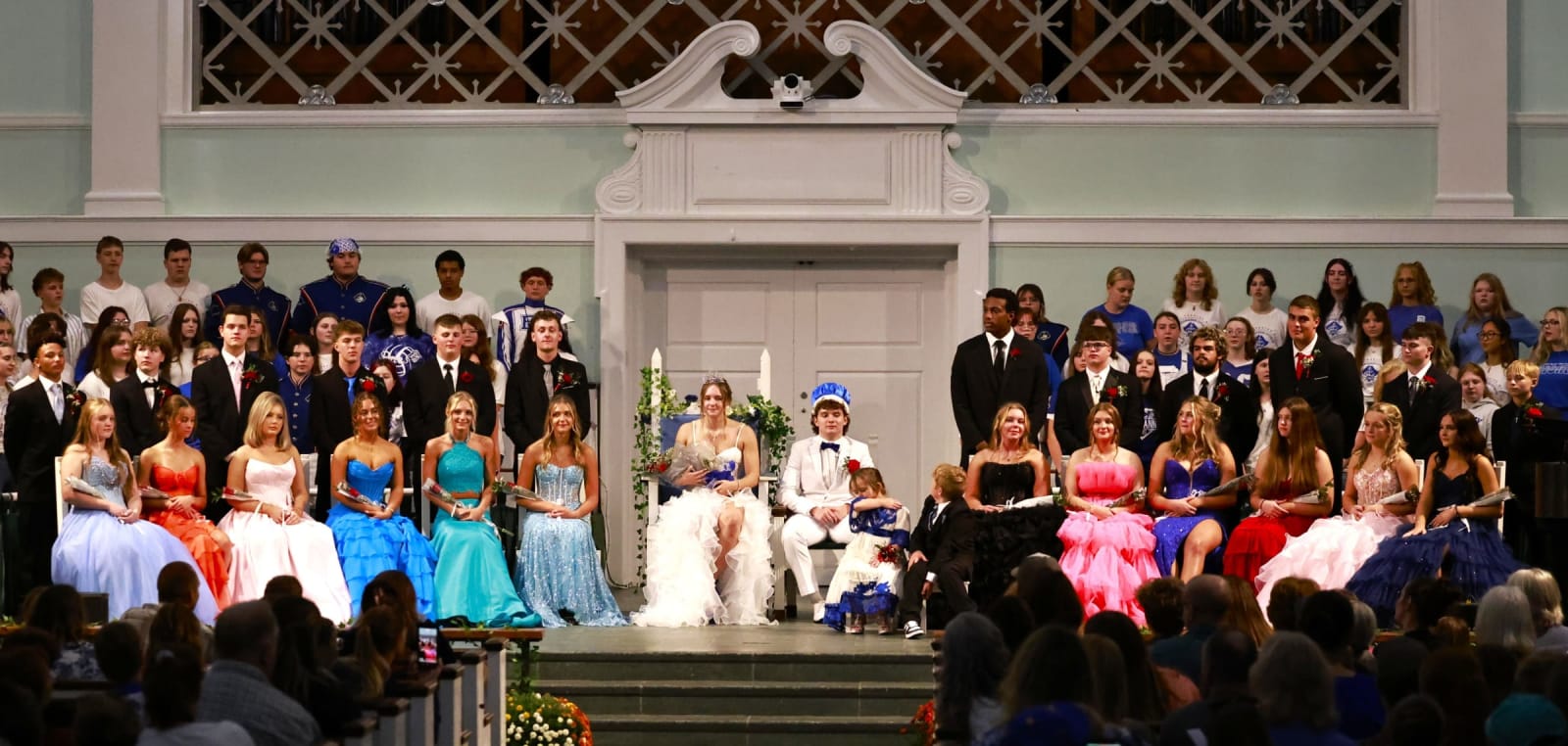 Homecoming court members sit and stand across the chapel stage with the audience in the foreground.