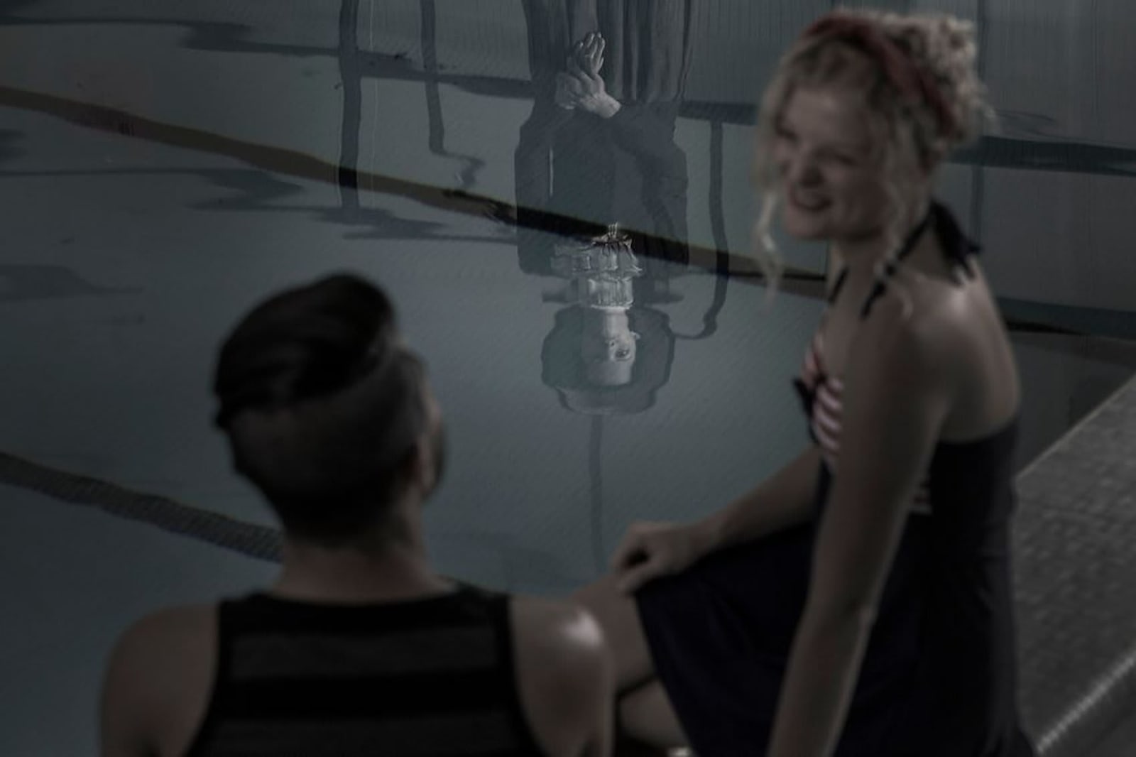 Two people sit by an indoor swimming pool, their reflections distorted in the water alongside the ghostly, pale face of a woman in historical clothing.